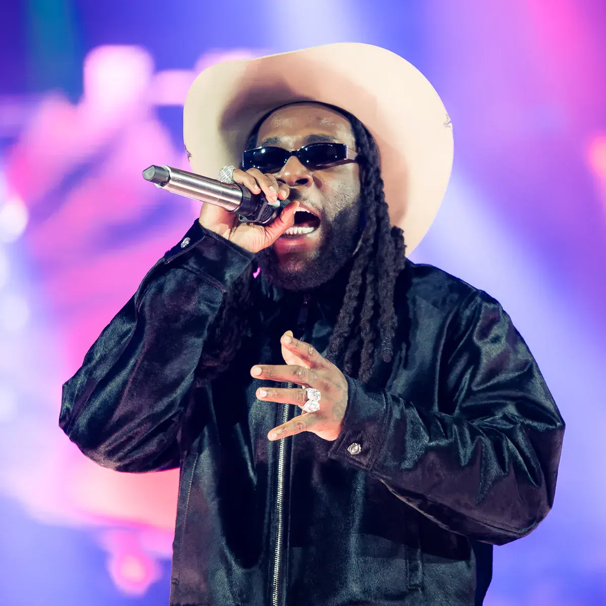 Burna Boy in a cowboy hat and dark glasses singing into a microphone under purple stage light