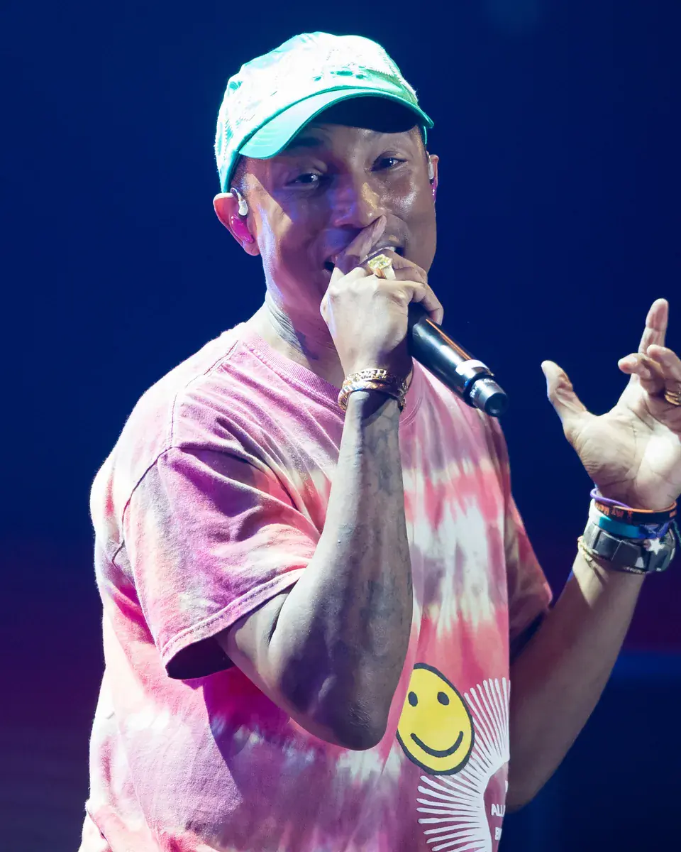 Pharrell Williams in a cap and a pink tie-dye shirt sings into a microphone on a blue-lit stage