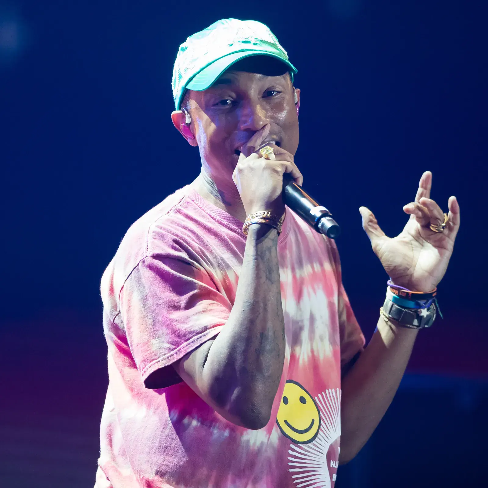 Pharrell Williams in a cap and a pink tie-dye shirt sings into a microphone on a blue-lit stage