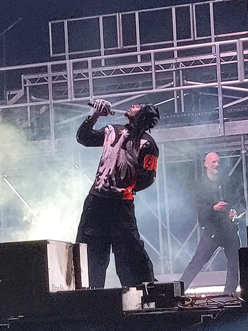 Playboi Carti in a black hood, microphone raised, standing on a stage monitor under scaffolding and smoke