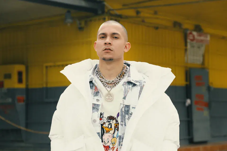 Tainy in a white puffer coat and chains, standing in a yellow-walled loading bay