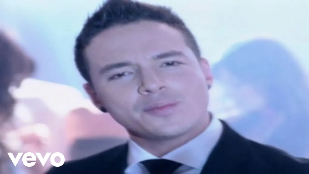 J Balvin in a dark suit and tie against a pale blue background, from the En lo oscuro video