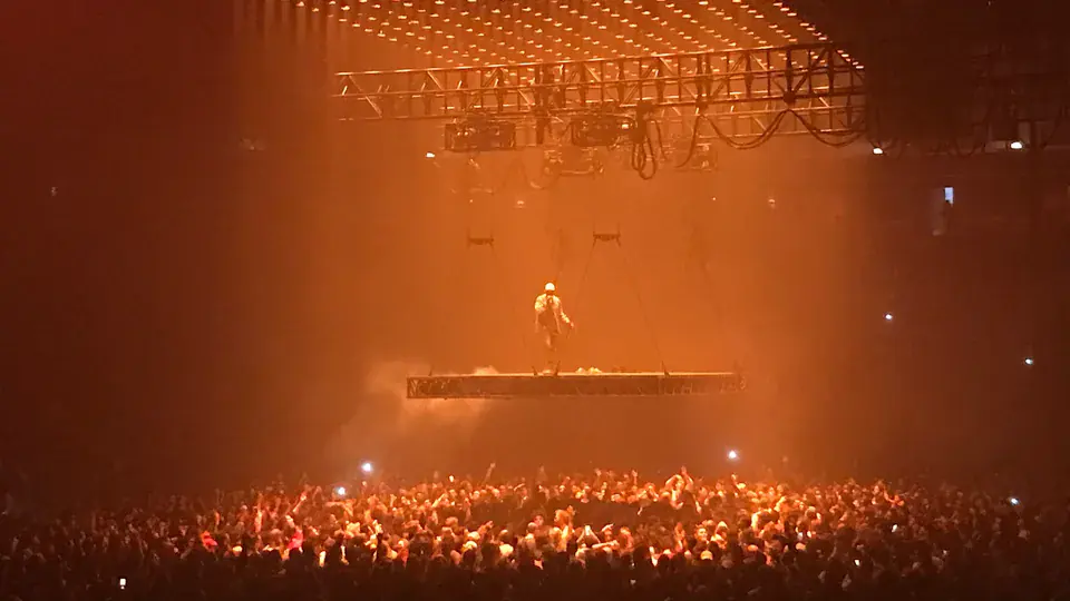 Kanye West on a stage suspended above a packed arena floor in orange light