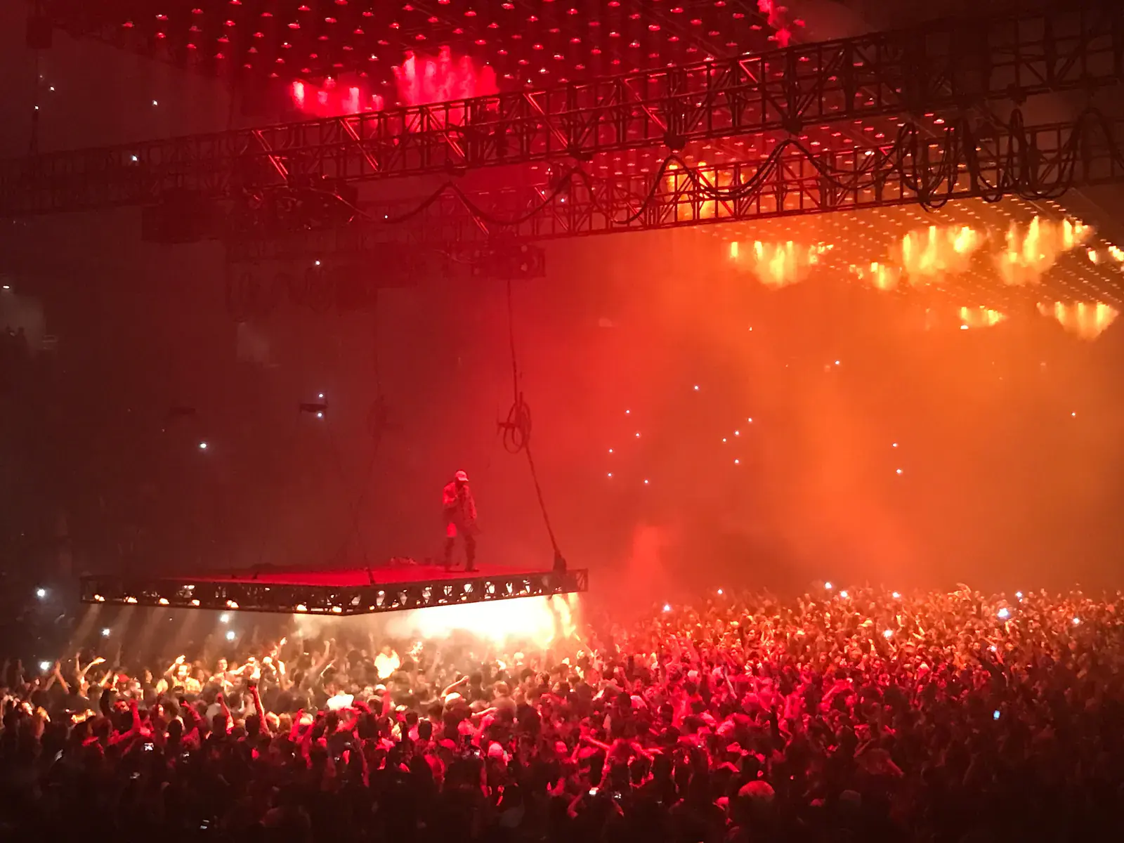 Kanye West stands on a stage suspended over a packed arena floor under rigs of red and orange lights